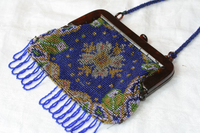 May include: Vintage beaded handbag with a dark brown frame and a beaded strap. The bag features a blue background with a floral design in gold, green, and white beads. Blue fringe hangs from the bottom.