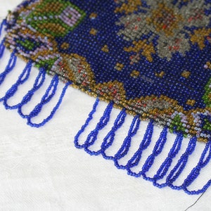 May include: A close-up of a beaded purse with a dark blue background and intricate floral patterns in shades of green, gold, and gray. The purse has a fringe of blue beads along the bottom edge.