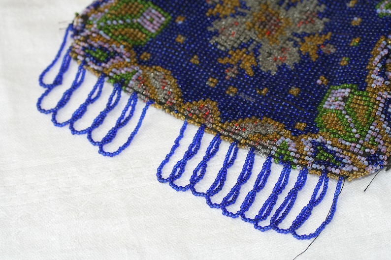 May include: A close-up of a beaded purse with a dark blue background and intricate floral patterns in shades of green, gold, and gray. The purse has a fringe of blue beads along the bottom edge.