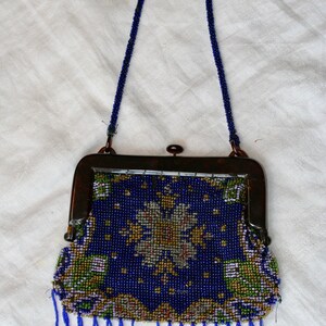 May include: Vintage beaded handbag with a dark blue background and a floral design in gold, green, and white. The purse has a dark metal frame, a beaded handle, and blue fringe along the bottom.