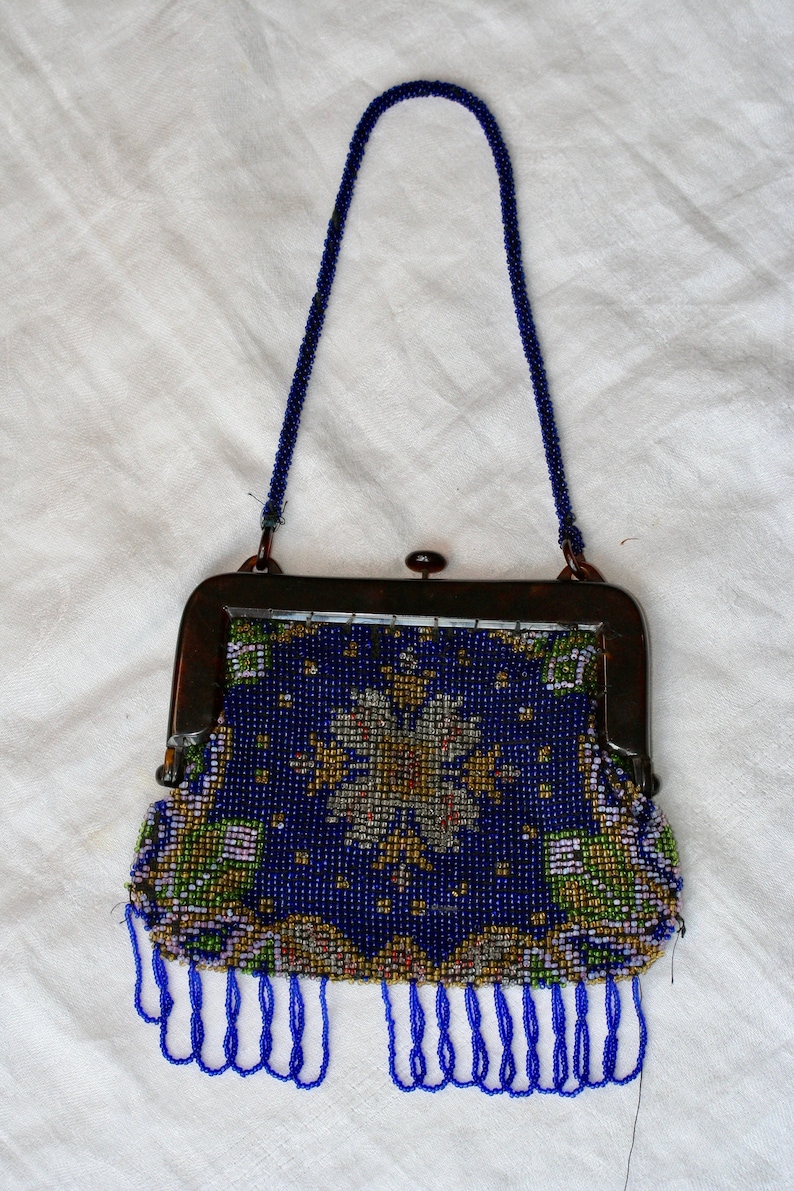 May include: Vintage beaded handbag with a dark blue background and a floral design in gold, green, and white. The purse has a dark metal frame, a beaded handle, and blue fringe along the bottom.