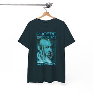 May include: A dark charcoal gray short-sleeve t-shirt featuring a cyan graphic print of Phoebe Bridgers. The text PHOEBE BRIDGERS is printed above the portrait, and the text LOST WEEKEND is printed below the portrait in a bold, sans-serif font.