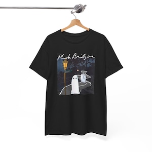 Könnte beinhalten: Ein schwarzes Kurzarm-T-Shirt mit einem Grafikdruck von drei weißen Geistern auf einer kurvigen Straße bei Nacht. Das Design zeigt eine gelbe Straßenlaterne, ein Straßenschild mit der Aufschrift Scott und den weißen Schriftzug Phoebe Bridgers.