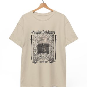 Puede incluir: Una camiseta beige de manga corta con un estampado gráfico en negro. El diseño incluye el texto Phoebe Bridgers y Punisher, una imagen central de una persona en un campo, bordes florales ornamentados, dos espadas verticales y dos aves.