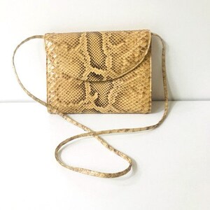 Könnte beinhalten: Eine kleine, hellbraune Clutch aus Schlangenleder mit Klappenverschluss und einem langen Riemen.