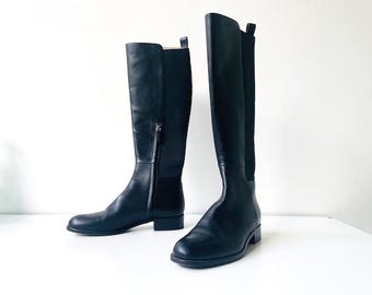 May include: A pair of black leather knee-high boots. One boot has a zipper on the side, and the other has an elastic panel. The boots are on a white surface.