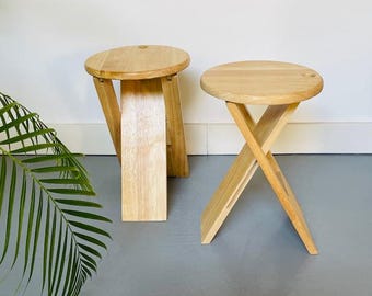 May include: Two wooden folding stools with round seats. The stools are made of light-colored wood and have a natural finish. One stool is folded, and the other is open, showing the crossed leg design. The stools are designed for indoor or outdoor use.