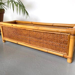 Peut inclure: Jardinière rectangulaire en bambou avec détails en canne tressée. La jardinière a une finition en bois naturel et est soutenue par quatre pieds courts. Un grand pot de plante est visible en arrière-plan.