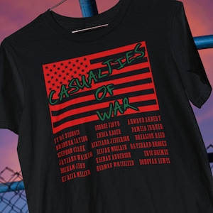 May include: Black t-shirt with a red, white, and blue American flag design. The flag has the words "Casualties of War" written in green on it. Below the flag is a list of names in green text.