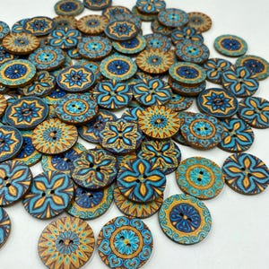 Puede incluir: Una colección de botones redondos de madera con varios patrones florales y geométricos intrincados en tonos azul, turquesa y dorado. Cada botón tiene dos agujeros en el centro para su fijación, con una superficie plana y un acabado impreso liso.