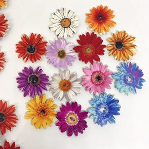 Puede incluir: Una colección de botones planos y redondos con forma de flores de margarita con dos agujeros centrales. Los botones presentan varios colores, incluyendo blanco, naranja, rojo, morado, rosa, amarillo y azul, con pétalos detallados y centros.