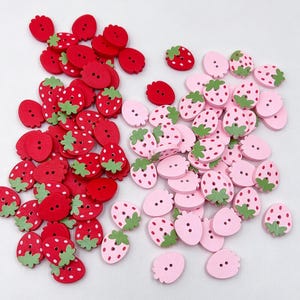 Puede incluir: Una colección de pequeños botones con forma de fresa en tonos rojo y rosa claro. Cada botón cuenta con dos orificios centrales para su fijación y un pequeño detalle de hoja verde en la parte superior. Los botones rojos tienen puntos blancos, mientras que los rosas tienen puntos rosa oscuro.
