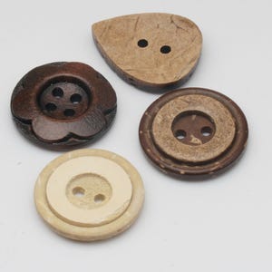 Pode incluir: Quatro botões de formas e cores variadas. Um botão castanho escuro em forma de flor com quatro furos. Um botão castanho claro em forma de gota com dois furos. Dois botões redondos, um castanho escuro com centro mais claro e dois furos, e um creme com dois furos.