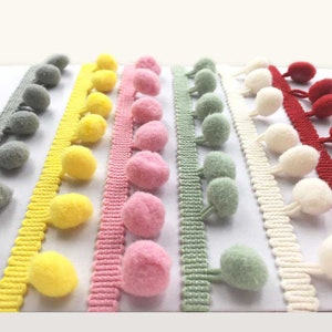 May include: A selection of pom-pom trim in a variety of colours, including grey, yellow, pink, green, white, and red. The trim is arranged in parallel lines, displaying the different colours and textures of the decorative trim.