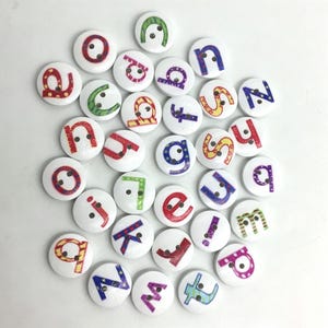 Puede incluir: Una colección de botones redondos blancos, cada uno con una letra minúscula colorida del alfabeto. Las letras presentan varios patrones, incluyendo rayas, puntos y estrellas, y cada botón contiene dos pequeños agujeros para coser.