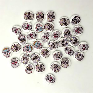 Puede incluir: Una colección de botones redondos blancos, cada uno con un diseño impreso de calavera de azúcar en colores. Las calaveras están decoradas con patrones intrincados en tonos morado, rosa, azul y negro, creando un aspecto uniforme en todas las piezas.
