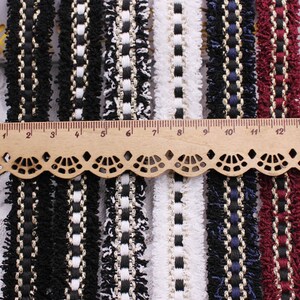 May include: Assortment of black, white, and red trim with fringe. The trim has a woven design with gold thread. The trim is measured against a ruler in inches.