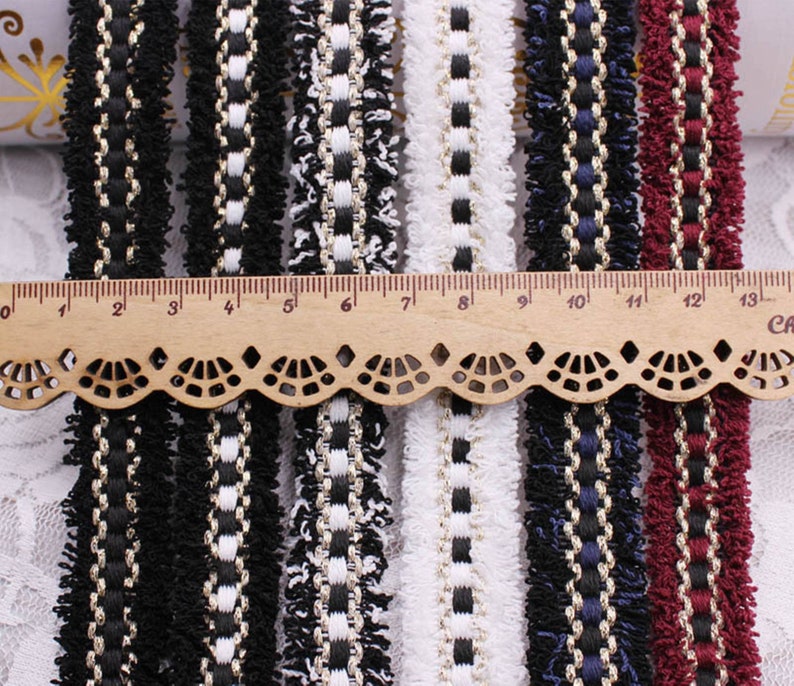 May include: Assortment of black, white, and red trim with fringe. The trim has a woven design with gold thread. The trim is measured against a ruler in inches.