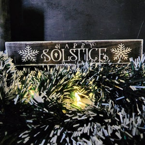 May include: A rustic wooden sign with the words "HAPPY SOLSTICE" in white lettering, flanked by snowflake designs. The sign is set against a backdrop of green and white tinsel with small lights.