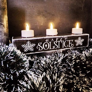 May include: A black wooden candle holder with three white LED candles. The holder has the words "HAPPY SOLSTICE" in white script, with snowflake designs. The holder is surrounded by black and white tinsel.