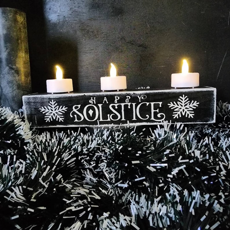 May include: A black wooden candle holder with the words "HAPPY SOLSTICE" in white script, flanked by snowflake designs. Three white LED candles with flickering flames sit atop the holder. The holder is surrounded by black and silver tinsel.