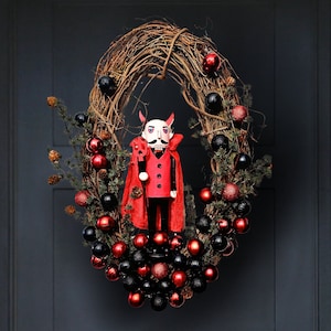 May include: A red and black Christmas wreath with a devil nutcracker in the center. The wreath is made of grapevine and is decorated with red and black ornaments.