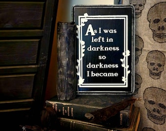 May include: A rectangular black wooden sign with white text that reads, "As I was left in darkness so darkness I became." The sign features a white border with a vine-like pattern and is positioned upright on a stack of old hardcover books.