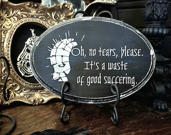 May include: An oval black wooden plaque with distressed white edges, featuring a white graphic of a grid-patterned head and the text: Oh, no tears, please. It's a waste of good suffering. The plaque is displayed on a black metal stand.