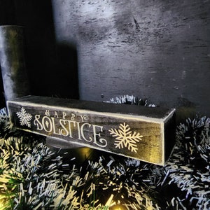 May include: A rectangular, wooden sign with the words "HAPPY SOLSTICE" in white script and snowflake designs. The sign is painted black with a distressed finish and sits on a decorative base with tinsel.