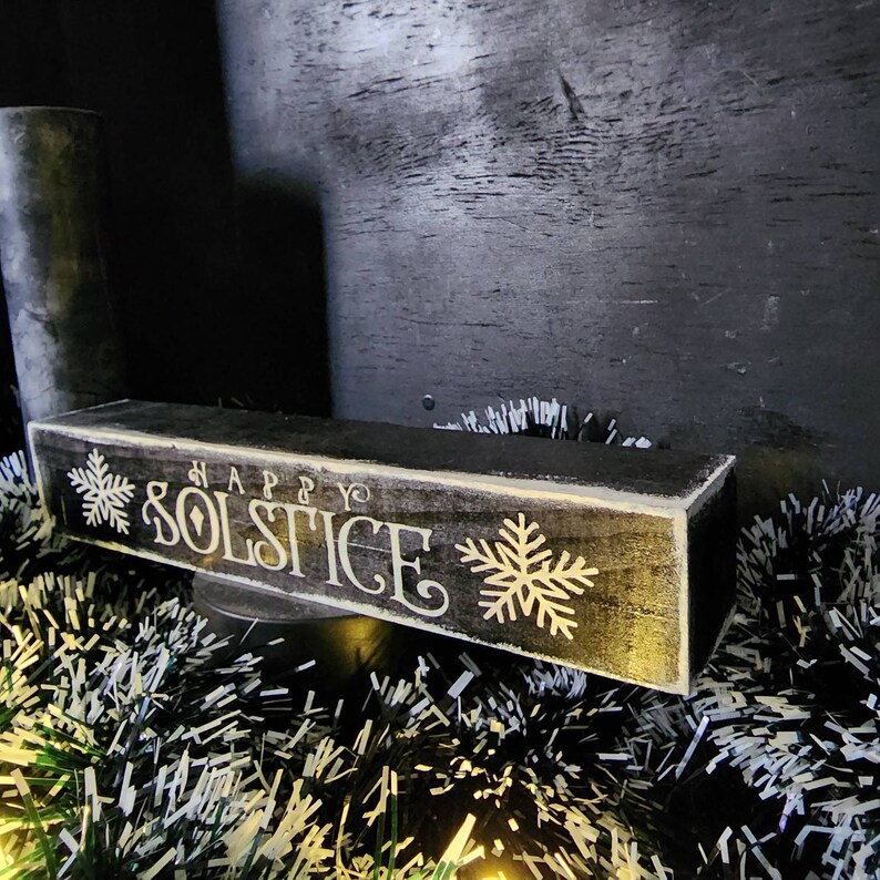 May include: A rectangular, wooden sign with the words "HAPPY SOLSTICE" in white script and snowflake designs. The sign is painted black with a distressed finish and sits on a decorative base with tinsel.