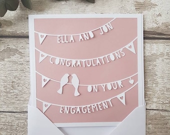 以下が含まれることがあります： ピンクのカードに、白い紙で切り抜かれた「エラとジョン 結婚おめでとう」と書かれたバナーがあります。バナーはハートと2羽の鳥で飾られています。