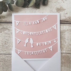 May include: A pink card with white paper cutouts of a banner that reads "Ella and Jon Congratulations on your Engagement". The banner is decorated with hearts and two birds.