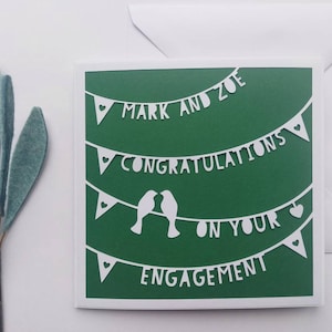 May include: A green card with white paper cutouts of a banner that reads "Mark and Zoe Congratulations on your Engagement". The banner is decorated with hearts and birds.