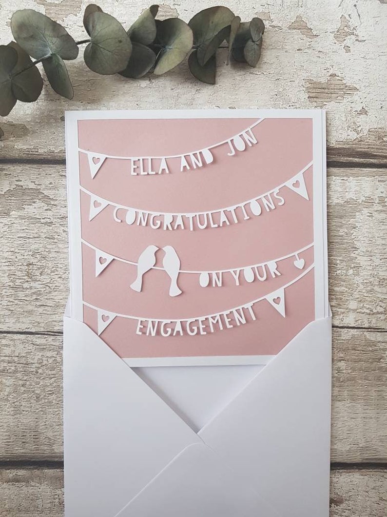 May include: A pink card with white paper cutouts of a banner that reads "Ella and Jon Congratulations on your Engagement". The banner is decorated with hearts and two birds.