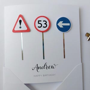 May include: A white card with three road signs. The signs are a red triangle with an exclamation point, a red circle with the number 53, and a blue circle with a white left arrow. The card has the name Andrew and the words Happy Birthday written on it.