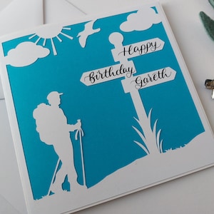 May include: A handmade birthday card with a hiker silhouette, sun, clouds, and a signpost that reads "Happy Birthday Gareth." The card features a white design on a turquoise background, with a white border. A unique, personalized gift.