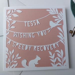 May include: A pink card with white cut-out designs of leaves, birds, and a squirrel. The card says "Tessa Wishing You A Speedy Recovery" in a banner design.