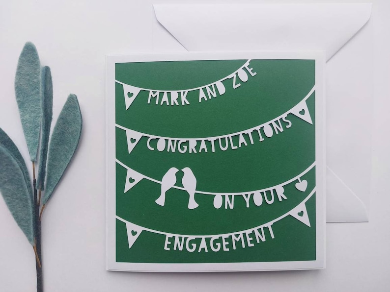 May include: A green card with white paper cutouts of a banner that reads "Mark and Zoe Congratulations on your Engagement". The banner is decorated with hearts and birds.