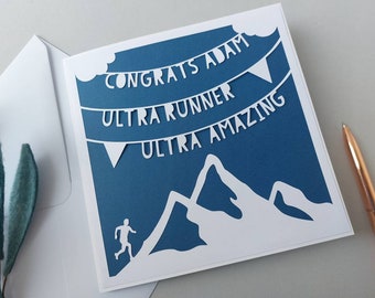 May include: A blue card with white cut-out text and a mountain scene. The text reads "Congrats Adam Ultra Runner Ultra Amazing".