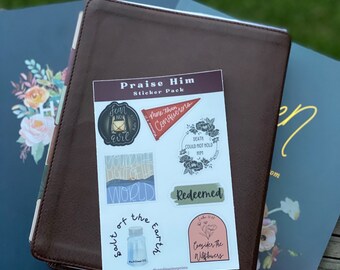 May include: A sticker pack with a brown leather-bound journal. The stickers feature religious quotes and illustrations, including a lantern, a jar of salt, a flower, and a banner with the words "More than Conquerors".