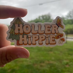 May include: A white sticker with the text "Holler Hippie" in orange and brown letters. The sticker has a green pine tree and a mushroom with a butterfly on the right side.