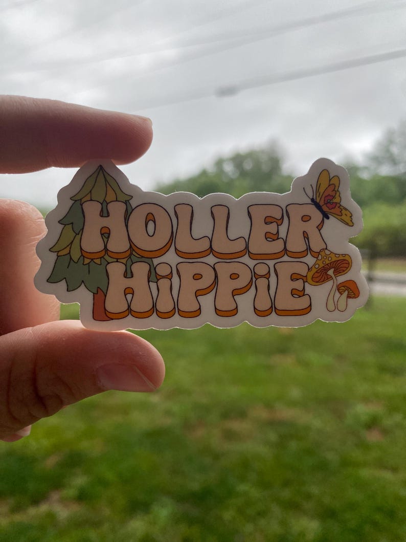 May include: A white sticker with the text "Holler Hippie" in orange and brown letters. The sticker has a green pine tree and a mushroom with a butterfly on the right side.