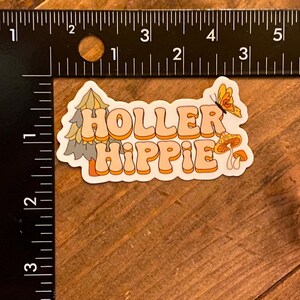 May include: A sticker with a retro design featuring the text "Holler Hippie" in orange and white letters. The sticker also includes a green pine tree, a yellow butterfly, and two orange mushrooms.