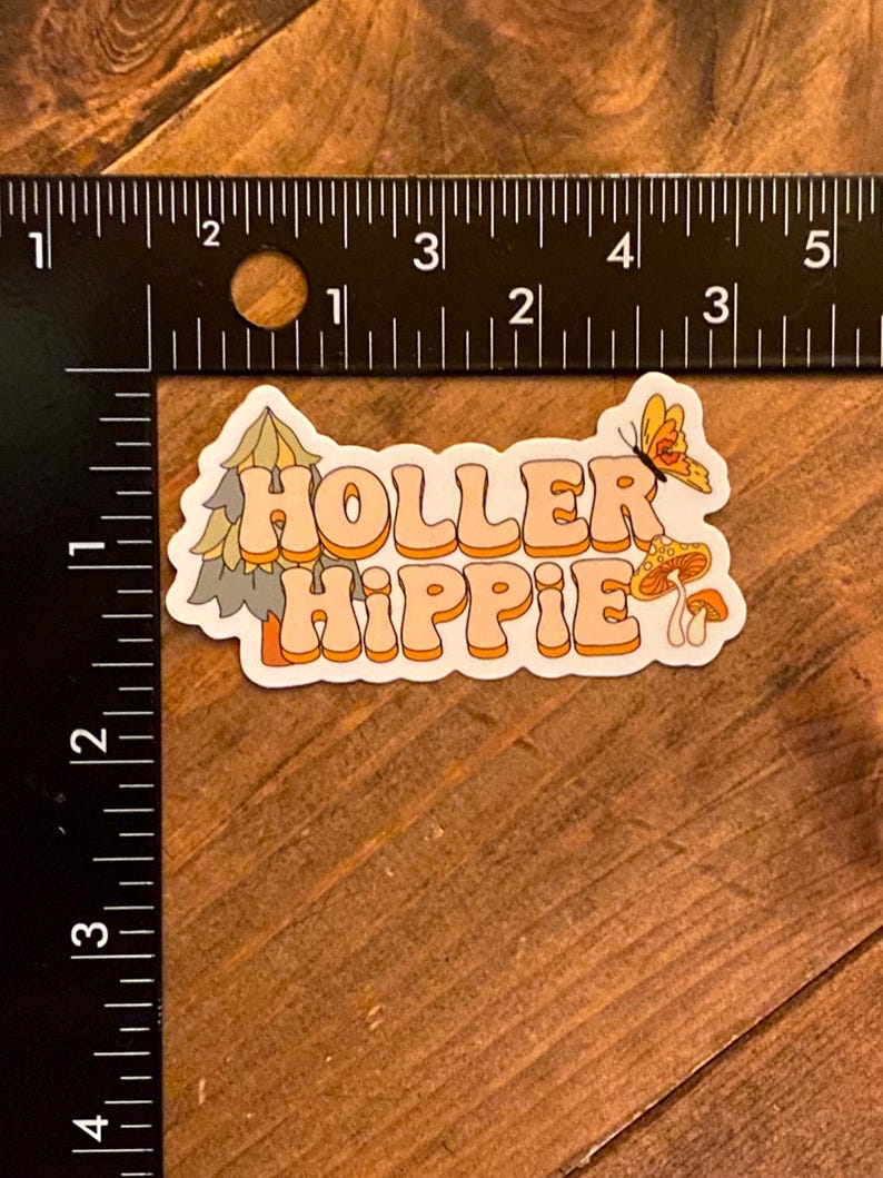 May include: A sticker with a retro design featuring the text "Holler Hippie" in orange and white letters. The sticker also includes a green pine tree, a yellow butterfly, and two orange mushrooms.