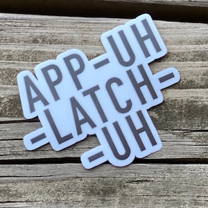 May include: A white sticker with black text that reads "App-Uh -Latch-Uh" on a wooden surface.