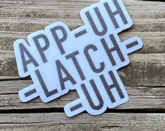 May include: A white sticker with black text that reads "App-Uh -Latch-Uh" on a wooden surface.