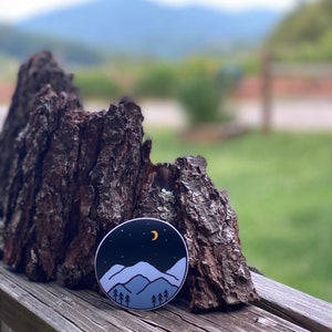 May include: A round sticker with a blue and white mountain range design against a black background with a crescent moon and stars. The sticker is on a piece of weathered wood.