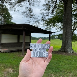 May include: A white sticker with black text that reads "Scholar from the Holler" held in a hand against a background of a wooden shelter and green grass.