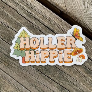May include: A white sticker with the text "Holler Hippie" in orange and peach letters. The sticker features a green pine tree, a yellow butterfly, and orange mushrooms.