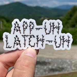 May include: A white sticker with the words "App-Uh Latch-Uh" written in black, with each letter formed by a stylized twig with leaves. The sticker is held in a hand against a blurry background of green hills and a blue sky.